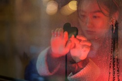 Korean girl think of lovely boyfriend drawing heart on cold fogged window covered with rain drops. Asian woman missing lover partner while being far on study or working. Love feelings and relationship