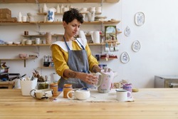 Artistic pottery master at work: happy woman in apron holding handmade jug working in ceramics studio. Craftswoman creating handicraft crockery in workshop. Craftsmanship and entrepreneurship concept