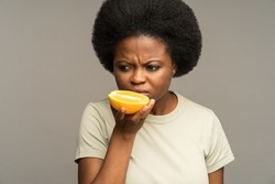 Unhappy African woman sniffing cut orange, suffering from anosmia during coronavirus disease, sick afro female has symptom of COVID-19 smell or taste loss, trying to smell food, isolated on grey