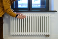 Cropped photo of woman warming hands near radiator at home after walking in cold winter weather, female touching barely warm battery during heating season, person near window checking heating system