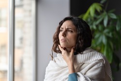 Upset sick woman touching painful swollen neck glands, suffering from throat infection, unhealthy female standing near window at home feeling pain with swallowing, selective focus