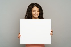 Excited young adult female hold blank placard looking down to empty copy space with pleasant smile. Beautiful millennial woman in pink outfit show while poster or banner isolated at studio background
