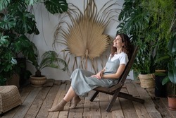 Mental health benefits of gardening. Young happy female gardener sitting in chair surrounded by various lush tropical plants. Smiling woman resting in home garden. Botanic boom and urban jungle trend