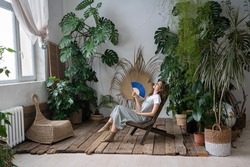 Stress relief after work from home. Relaxed young lady spend time in cozy indoor garden with monstera plant sitting with closed eyes and blowing fan. Love for plants, wellness and wellbeing concept