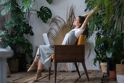 Work-life balance. Happy female freelancer with closed eyes relaxing while working remotely in home garden full of exotic plants. Young woman resting during remote work at urban jungle home office