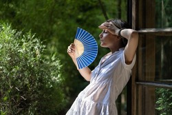 Young woman suffering heatstroke outdoors using fan to get fresh air. Unhappy girl feel bad of hot temperature outside. Frustrated female tired of summer season heat in fatigue touch forehead