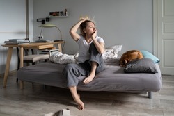 Sad middle-aged female sits on bed near dog and looks at window with absence of starting day. Thoughtful worried Scandinavian woman feels depressed and lonely. Shabby woman thinks about difficult