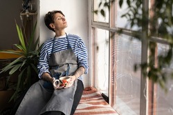 Tired female artisan with cup of tea in hands on windowsill, resting in ceramics studio, dreaming to open her own pottery studio. Peaceful female ceramist taking break during workday in workshop