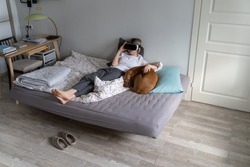 Lonely middle-aged woman wearing VR glasses lying on unmade bed with pillows and hugs vizsla dog. Mature female in pyjama plays interesting games avoiding problems and difficulties in life. Addiction.