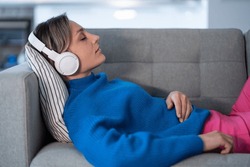Young woman wearing wireless headphones and blue sweater lies on comfortable sofa and enjoys listening to calm music. Female rests from long day working as freelancer at home office closeup