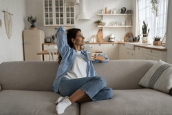Happy dreamy young woman wearing casual clothes resting with cup of tea at cozy home, smiling female sitting on couch enjoying favorite hot drink, sipping it slowly to relax. Hygge and slow life