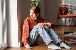 Upset teen girl sit on floor sadly look out window worried about teenage problem at school and communication with parent. Worried girl tensely suffer about bullying at school, unrequited love with boy