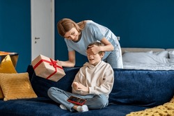 Young happy loving mother closing eyes of child congratulating little boy son with happy birthday at home, smiling mom giving wrapped gift box to kid sitting on sofa with smartphone. 