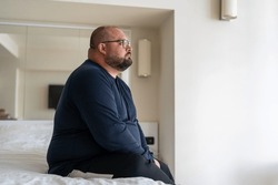 Sad fat lonely man sitting on bed in hotel and looking out window, thinking about life problems, no strength change, side view overweight depressed man no friends and mental disorders