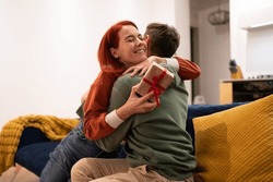 Overjoyed happy young woman holding gift box hugging husband feeling excited getting present from boyfriend. Family couple celebrating birthday and anniversary together at home, sitting on sofa