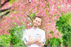 A guy with a beard embraces himself and smiles in the park near the flowering trees