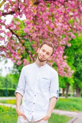 A guy with a beard holds his hands in his pockets and looks at the camera lens in a park near the flowering trees.