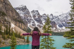 Back view of young woman, with open arms, at canadian landscape, with turquoise water lake, pine trees and Rocky Mountains on background, in Lake Moraine, Banff National Park, Alberta, Canada.