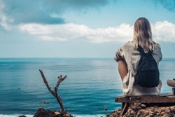 girl with blond hair sits hugging her legs on a cliff,incredible view of the ocean,on the model gray jumper and black backpack, she looks into the distance, endless horizon,life in travel, tourism