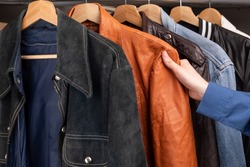 Man choosing clothing in a second hand store. Various vintage suede leather and jeans jackets hang on clothing rack. Thrifting and sustainability in clothing concept