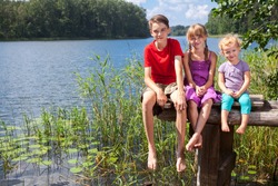 Three children of different age - teenager boy, elementary age girl and toddler girl sitting on a wooden pier by a forest lake looking at camera smiling