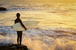 Surfer stands with a board on the Atlantic Ocean coast at sunset and waits for a suitable wave to start riding on a board and enjoy