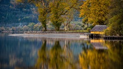 A small yellow boathouse in the Trossachs National park reflecting in the calm water of Loch Ard