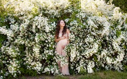 Young pregnant woman in a spring garden. Caucasian woman with long brown hairs in long romantic dress near the blooming spirea white bush. Concept of new life of nature and human. Waiting of baby