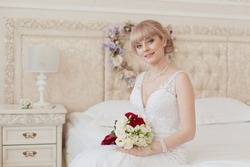 Pretty young Bride in elegant wedding dress sitting on a bed. Blonde-haired woman with wedding hair-style in royal room of hotel. Bride's morning in light classic interior. Wedding day ceremony