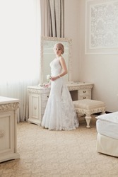 Bride's morning in light classic interior. Pretty young Bride in elegant wedding dress near a window. Blonde-haired woman with wedding hair-style in royal room of hotel. Wedding day ceremony