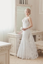 Bride's morning in light classic interior. Pretty young Bride in elegant wedding dress near a window. Blonde-haired woman with wedding hair-style in royal room of hotel. Wedding day ceremony