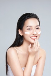 Beauty portrait image of pretty asian woman smiling and applying face cream isolated over light grey background