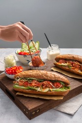 Banh Mi - Vietnamese sandwich with sausage, pork, lettuce, tomato and arugula on the light grey texture background. With hand and chopstick to make photo look natural