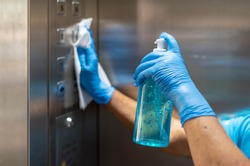 Hand in blue surgical gloves using disinfectant from the bottle spraying an elevator push button control panel.Disinfection,cleanliness and healthcare,Anti bacterial and Corona virus (COVID-19). 
