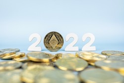 Ethereum 2022 price prediction concept. Ether coin standing next to cryptocurrency tokens and year numbers on blue background. Close-up, soft focus.