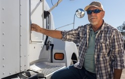 Hispanic middle aged semi truck driver sits next to truck. Experienced senior trucker next to big rig at the job. Portrait of transportation and logistics worker next to equipment on a sunny day.