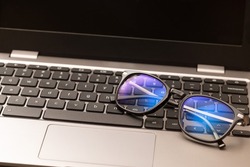 Reading glasses with coating filter to prevent Computer Vision Syndrome (CVS) sit on laptop keyboard. Concept of digital eye strain caused by screens and monitors of smartphones and computer devices.