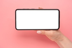 Hand holding mobile phone in horizontal position for mockup. watching streaming video on cellphone on pink table background. Close up of horizontal black smartphone with blank screen in woman hands