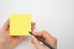 Hand written notes black pencil on yellow sticker. white table background. woman hand writing on yellow sticky notes. female hand holding note paper and making note