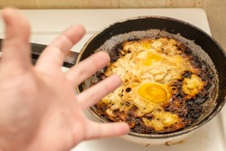 view of overcooked, burnt eggs, omelet on an old cast iron frying pan on an electric stove against the background of the hand of an emotionally outraged person