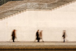Motion blurred business people walking in front of a bright concrete wall.