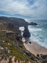 View of sharp cliffs and small sand beach with ocean waves, rocks, stones and green vegetation at wild Rota Vicentina coast near Vila Nova de Milfontes, Portugal.