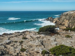 View sea shore with ocean waves, sharp rocks and stones and green bush at Rota Vicentina wild coast near Vila Nova de Milfontes, Portugal.
