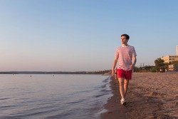 A handsome, young guy, a man in a white T-shirt with stripes and shorts is walking along the embankment of the river, the sea. Walking at the resort. Rest by the beach.