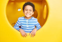 Portrait of cute African-American boy smiling happily peeking from round window of yellow cheese like box having fun on playground, copy space