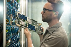 Serious skilled young bearded server installation specialist in glasses holding circuit board and examining server racks while working in datacenter room