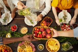 Top view at multi-ethnic group of people enjoying dinner together during outdoor Summer party, focus on hands handing fresh fruits and berries across wooden table, copy space
