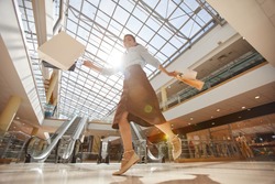 Warm-toned low angle portrait of happy young woman jumping in mall and holding shopping bags, copy space