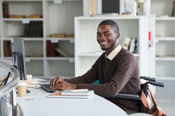 Portrait of disabled African-American man using computer and smiling at camera while studying in college library, copy space
