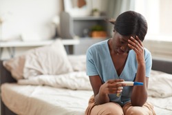 Portrait of worried African-American woman looking at pregnancy test while sitting on bed at home, copy space